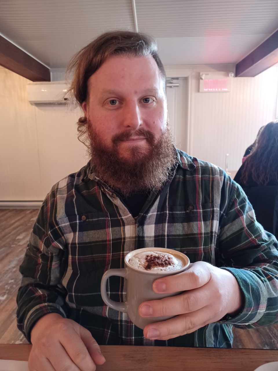 Un homme, assis à une table, regarde l'objectif tout en tenant un café au lait souppoudré de cacao. L'homme porte une barbe rousse et bien garnie ainsi qu'un chemise à carreau. 